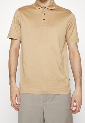 Mann trägt ein beiges Kurzarm-Poloshirt mit drei Knöpfen und hellgraue Hose, steht mit entspannt an den Seiten hängenden Armen.