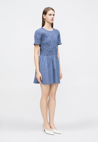 Femme debout en mini-robe à carreaux bleue à manches courtes et talons blancs, sur un fond uni clair.