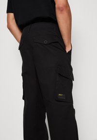 Schwarze Cargo-Hosen mit lockerer Passform, die über Seitentaschen, einen Knopfverschluss und ein kleines Markenetikett auf der Gesäßtasche verfügen.