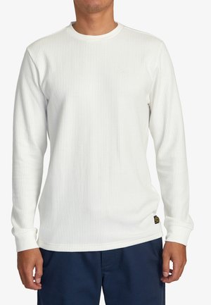 Chemise blanche à manches longues en rib d'un col rond, avec des poignets ajustés et un petit logo près de l'ourlet, portée sur un pantalon sombre.