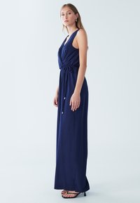 Mornarsko modra maxi obleka brez rokavov z globokim V-izrezom, poudarjenim pasom in detajlom z vrvico. Gladek material z udobnim krojem; kombinirano s črnimi petami.