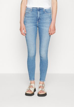 Slim-fit lyseblå jeans iført hvide sandaletter med åbne tå-dele og sølv spænder på en person, der står mod en ensfarvet hvid baggrund.