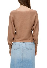 Frau, die einen gerippten, langärmeligen braunen Pullover und einen hellblauen Jeansrock trägt, von hinten vor einem weißen Hintergrund gezeigt.