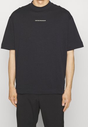 Person trägt ein schwarzes oversized Emporio Armani T-Shirt mit kurzen Ärmeln und kleinem weißem Logo-Schriftzug auf der Brust.