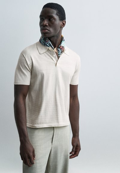 Homme à la peau foncée portant un polo à manches courtes crème, un pantalon gris clair et une écharpe multicolore à motifs autour du cou, regardant vers la gauche.