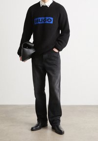 Schwarzer gerippter Pullover mit blauem "HUGO"-Logo, kombiniert mit dunklen Jeans und schwarzen Lederschuhen. Halte eine schwarze, strukturierten Handtasche.