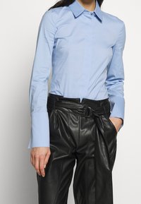 Light blue long-sleeve shirt with a pointed collar and button closure, paired with high-waisted black leather trousers featuring a belt.