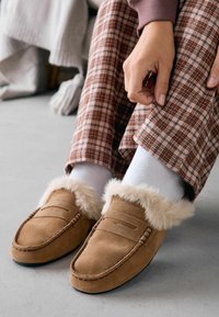 Chaussons en daim marron avec une doublure en fourrure, décorés de détails de couture et d'une semelle plate, portés avec un pantalon à carreaux et des chaussettes blanches.