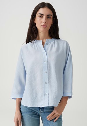 Vrouw met lang donker haar die een lichtblauw blouse met knopen en driekwart mouwen en blauwe jeans draagt, staand tegen een effen achtergrond.