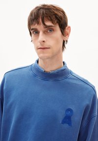 Blaues Sweatshirt mit hohem Kragen, aus weichem Stoff. Verfügt über ein strukturiertes blaues Logo auf der Brust und sichtbare Nähte.