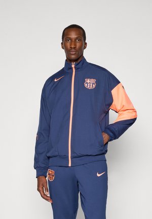 Navy tracksuit jacket with orange accents, featuring a high collar, zipper closure, and Barcelona logo on chest and sleeve. Smooth texture.