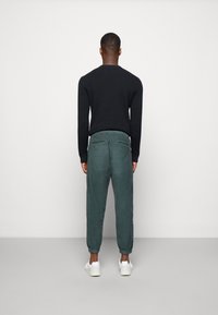 Černý pletený svetr a zelené zúžené kalhoty s elastickými manžetami, doplněné bílými teniskami, zobrazené zezadu na jednobarevném pozadí.