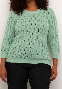Pull en tricot vert menthe avec travail en filigrane texturé, présentant un motif ondulé et des manches trois-quarts, fini par un ourlet côtelé.