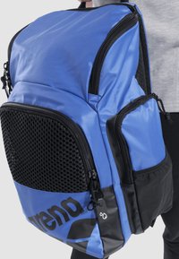 Mochila azul hecha de un tejido duradero con un bolsillo frontal de malla negra, múltiples compartimentos con cremallera y una base impermeable. Diseño del logo en negro.
