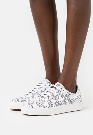 Fødder iført hvide sneakers med lav top, sorte abstrakte linjemønstre og hvide snørebånd på en hvid baggrund.