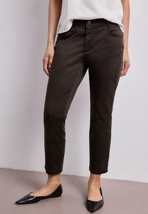 Kvinde iført sorte slim-fit ankellange bukser, hvid top og sorte spidse flade sko, stående mod en ensfarvet lys baggrund.