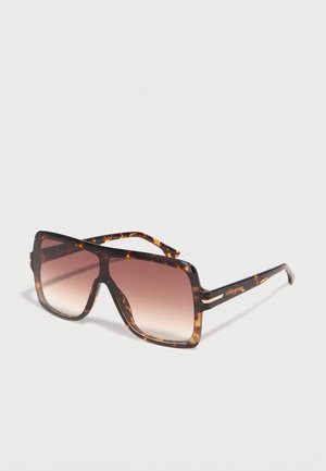 Nadmerné štvorcové slnečné okuliare s rámom v korytnačinovej farbe a hnedými šošovkami s prechodovým efektom, naklonené na bielom povrchu.