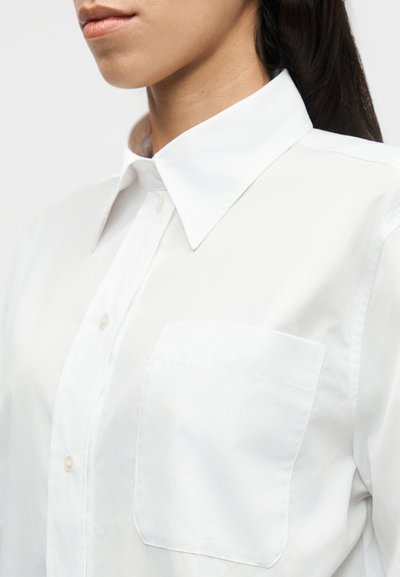 Λευκό πουκάμισο με κουμπιά και γιακά, με μία τσέπη στο στήθος. Το ύφασμα φαίνεται λείο και ελαφρύ, με καθαρό, κομψό σχέδιο.