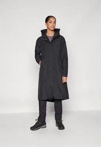 Fekete vízálló hosszú kabát kapucnival, cipzáras záródással és oldalzsebekkel; sötét nadrágra és masszív fekete csizmára viselve. Sima textúra.