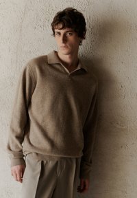 Pulover tricotat bej cu guler cu plachetă pe față, manșete și margine ribbed. Purtați peste pantaloni bej deschis, în fața unui perete texturat.