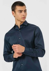 Camicia blu navy con bottoni, realizzata in tessuto morbido, dotata di colletto a punta, maniche lunghe e polsini con bottoni. Esterno con un leggero lucido.