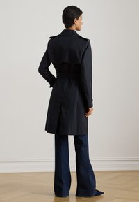 Trench-coat noir avec un devant croisé, une ceinture cintrée et un dos évasé. Tissu lisse avec des poignets boutonnés et une fente arrière.