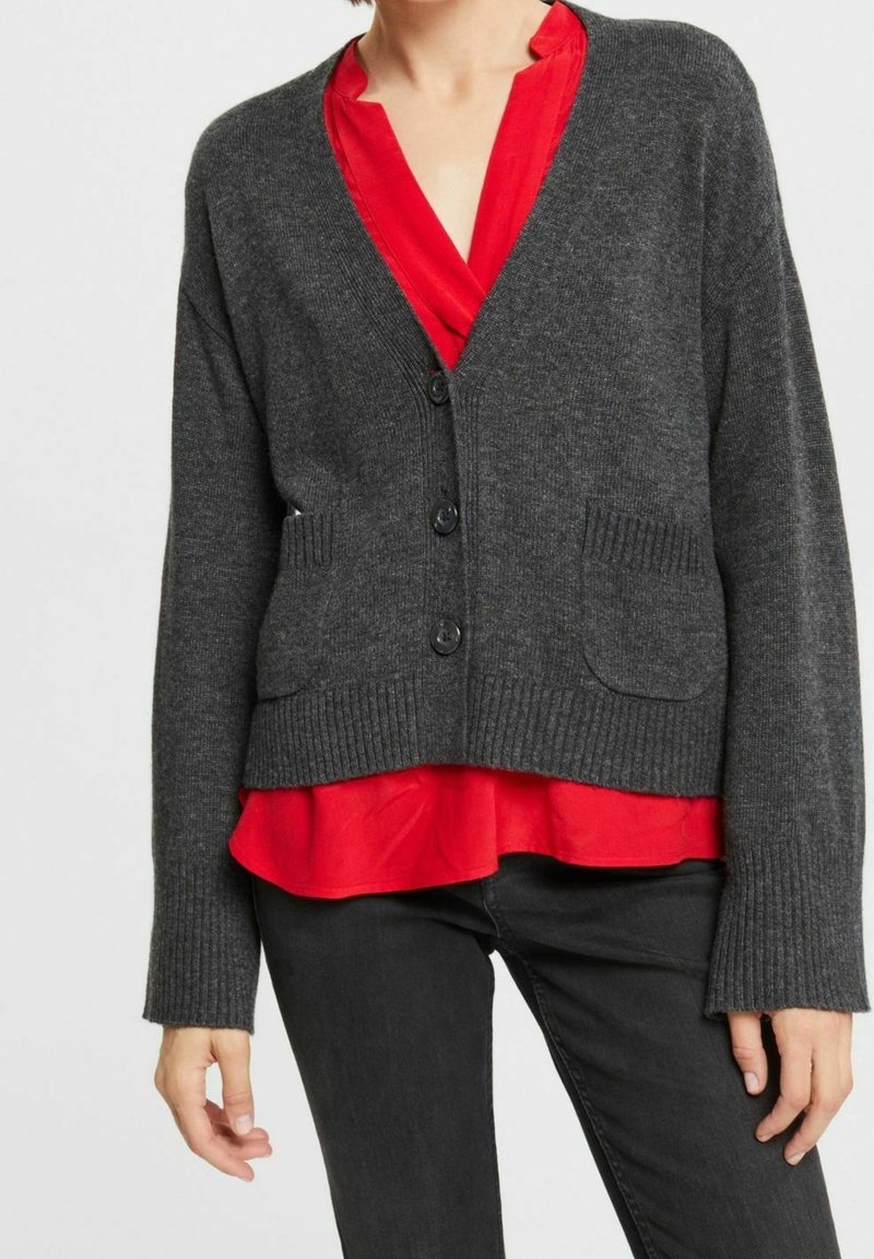 Kohlegrauer, gestrickter Cardigan mit gerippten Bündchen und Saum, der zwei Taschen auf der Vorderseite und fünf dunkle Knöpfe hat, über einer roten Bluse getragen.