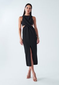 Čierne šaty bez rukávov so skrúteným predným dizajnom a rozparkom až po stehná. Doplnkové zlaté sandále na remienky. Jednoduché a elegantné.