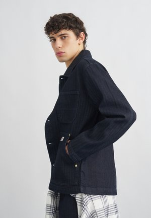 Junger Mann mit lockigem Haar, der eine dunkle Jeansjacke, Ohrringe und einen karrierten Rock trägt und vor einem schlichten hellen Hintergrund steht.