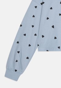 Lyseblå, kortermet langermet skjorte med svarte hjerte mønstre. Myk stoff med samlet ermer og en avslappet passform. Enkel utringning.