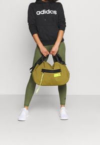 Personne portant un sweat à capuche Adidas noir, des leggings verts et des baskets blanches tenant un sac de sport Adidas vert olive avec des poignées noires.