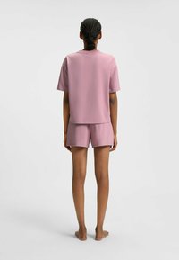 Persona con capelli intrecciati che indossa una maglietta rosa a maniche corte larga e pantaloni corti abbinati, in piedi scalza su uno sfondo bianco semplice.