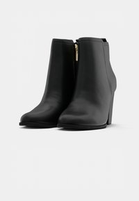 Bottines en cuir noir avec un talon haut, arborant un design élégant, une texture lisse et un détail de zip doré sur le côté.