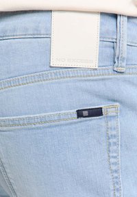 Lichtblauwe denim jeans met een witte leren patch met het label "NO EXCESS." Heeft contrasterende stiksels en een kleine marineblauwe accentzak.