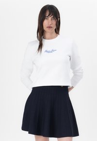 Felpa bianca a maniche lunghe, con logo blu "Guess Jeans". Abbinata a una gonna plissettata blu navy con dettagli di bottoni.