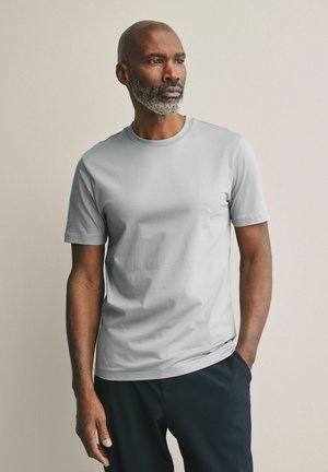 Camiseta de algodón gris con cuello redondo, mangas cortas, textura suave y un corte relajado, combinada con pantalones oscuros. Diseño simple sin patrones.