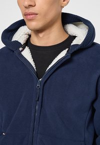 Jongere in een marineblauwe fleece hoodie met witte sherpa voering, gouden ringoorbel en een zwart crewneck shirt eronder.