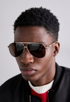 Des lunettes de soleil marron avec des accents dorés et une rayure verte-rouge-verte sur la branche, portées avec une veste bomber noire sur un pull rouge et blanc.