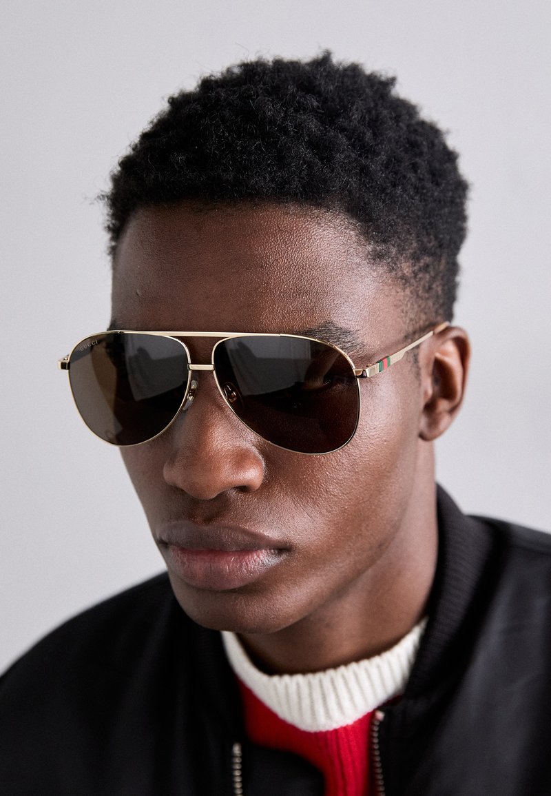 Hnědé sluneční brýle se zlatými akcenty a zeleno-červeno-zeleným pruhem na spánku, nošeny s černou bomber bundou přes červený a bílý svetr.