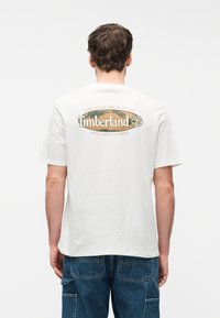 Άνδρας που φοράει λευκό t-shirt Timberland με το λογότυπο και το κείμενο «Αέρας, Νερό, Γη και Ουρανός» στην πλάτη, συνδυασμένο με μπλε τζιν παντελόνι καμπαρντίνα.