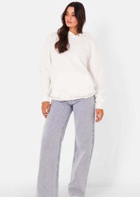 Vrouw draagt een wit hoodie met zak aan de voorkant en lichtblauwe wijd uitlopende jeans, staand tegen een effen witte achtergrond.