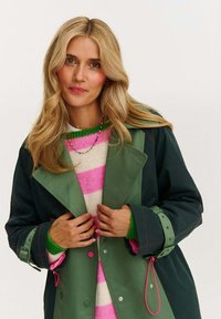 Zelený voděodolný kabát s růžovými akcenty, nošený přes růžovo-bíle pruhovaný svetr. Vybaven širokým límcem a nastavitelnými manžetami.