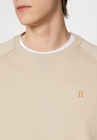 Sudadera beige con cuello y yugo acanalados. Presenta un acento bordado en naranja con "II", textura suave y un diseño clásico de cuello redondo.