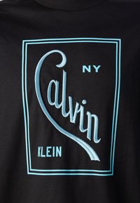 Juodas medvilnės marškinėliai su šviesiai mėlynu išsiuvinėtu logotipu "Calvin Klein", stilizuotais rašmenimis, esančiais stačiakampio rėme.