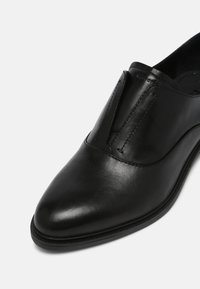 Chaussure de ville en cuir noir à enfiler avec un bout pointu et des panneaux élastiques sur les côtés, sur fond blanc.