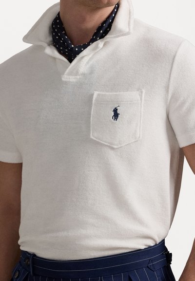 Polo shirt blanc texturé avec un col demi et une poche sur la poitrine. Il présente un logo brodé bleu marine. Porté avec un foulard de cou bleu à motifs.