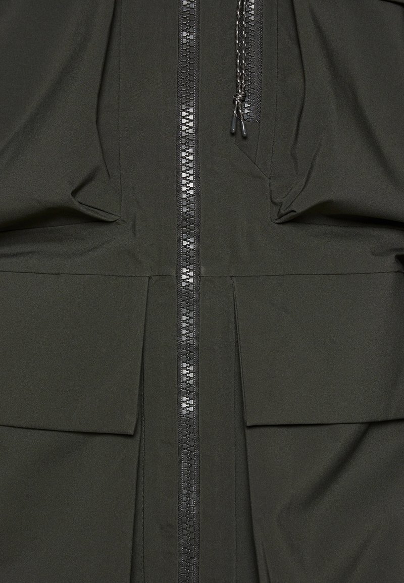 Chaqueta verde oscuro con una textura de tela suave, que cuenta con un cierre central, dos grandes bolsillos frontales y un diseño con plisados en el dobladillo.