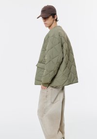 Oliven grøn quiltet jakke med løs pasform og ribstrik i ærmerne, parret med lyst beige løstsiddende jeans. Brun kasket med teksten "DETROIT".