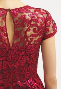 Robe en sequins rouges avec des motifs floraux complexes, un dos transparent, une fermeture à petit bouton et des manches courtes. Design texturé et ajusté.