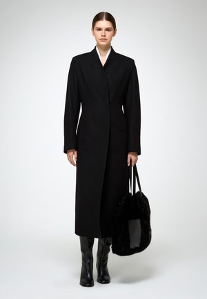Cappotto lungo nero con taglio dritto, colletto alto e chiusura frontale. Abbinato a stivali neri al ginocchio e una morbida borsa nera.